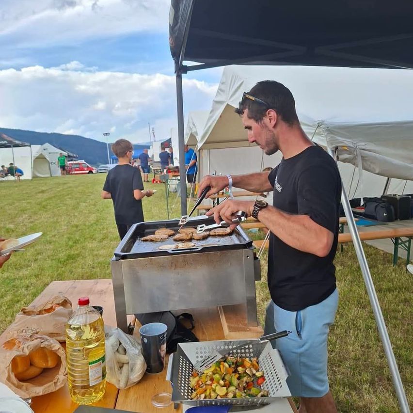 Ein Mann kocht Essen auf einem Grill unter einem Zelt bei einer Outdoor-Veranstaltung, während ein Junge zuschaut.