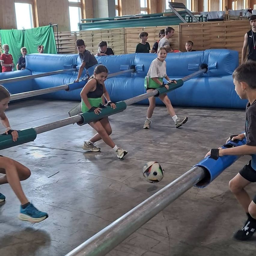 Mehrere Kinder spielen in einer Turnhalle ein Spiel mit großen Stangen und einem Fußball. Einige laufen, während andere die Stangen halten.