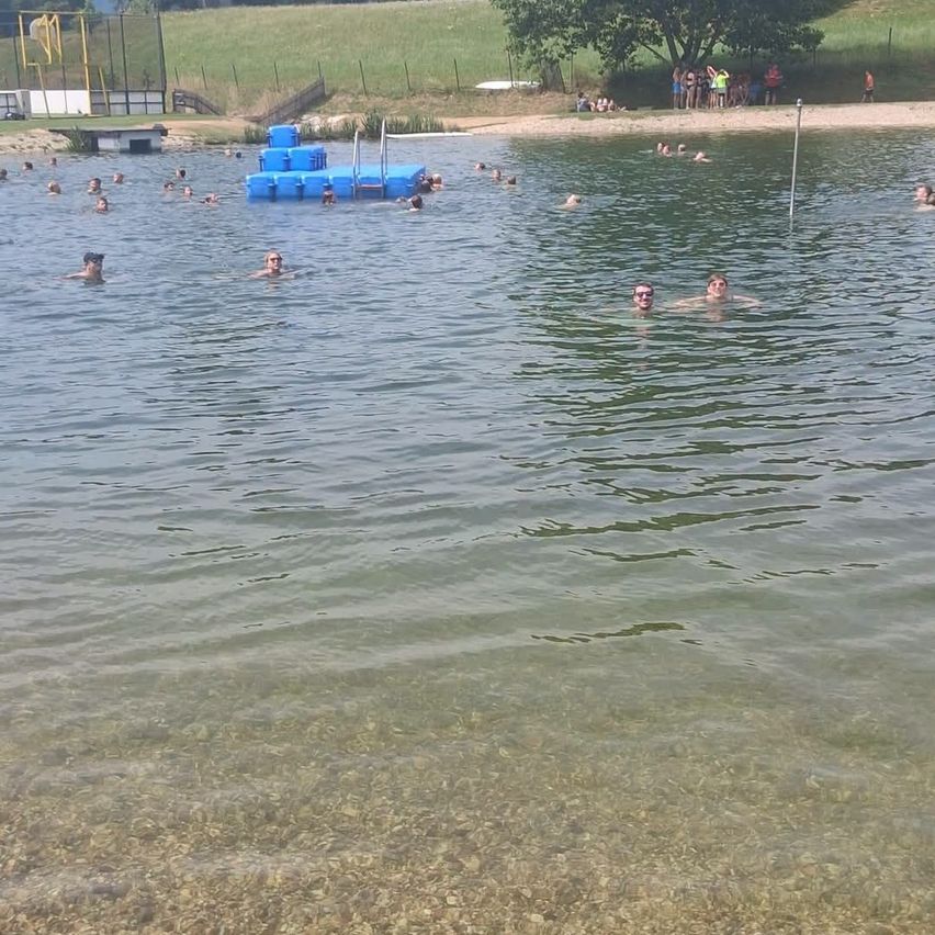 Eine Gruppe von Menschen schwimmt in einem See mit einer blauen schwimmenden Plattform in der Nähe. Einige stehen am Ufer.