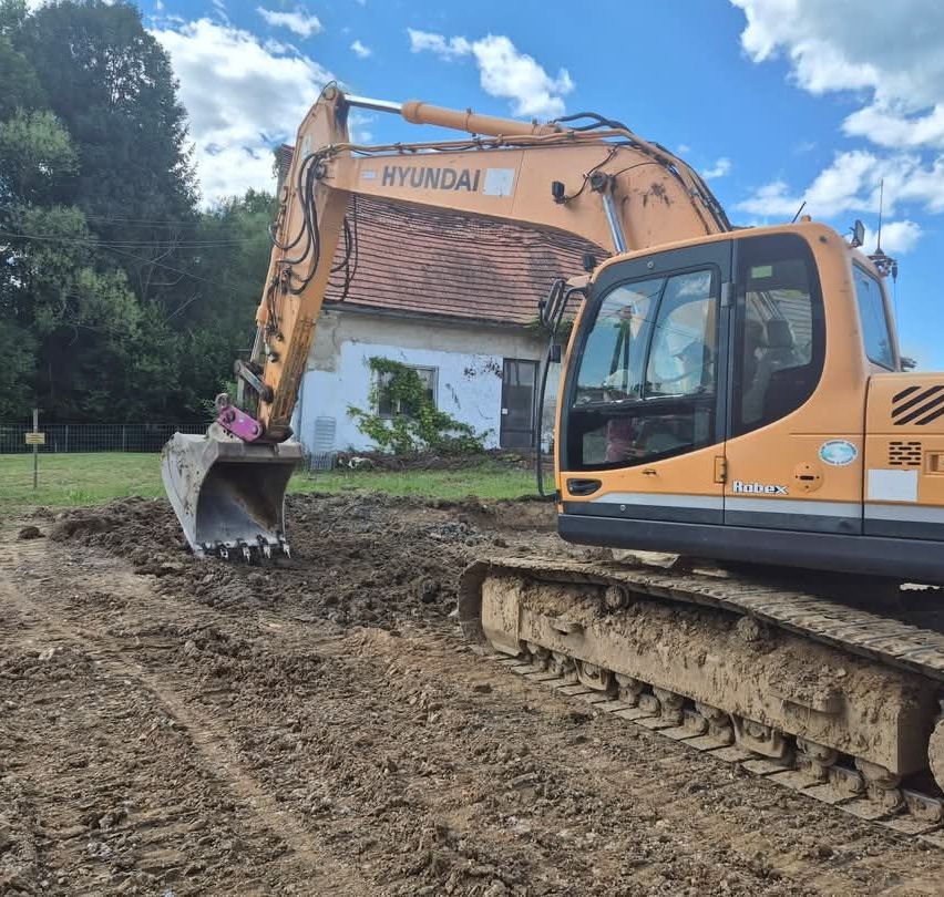 Ein Hyundai-Bagger steht auf einer schlammigen Fläche, mit einem Haus im Hintergrund unter einem teilweise bewölkten Himmel.