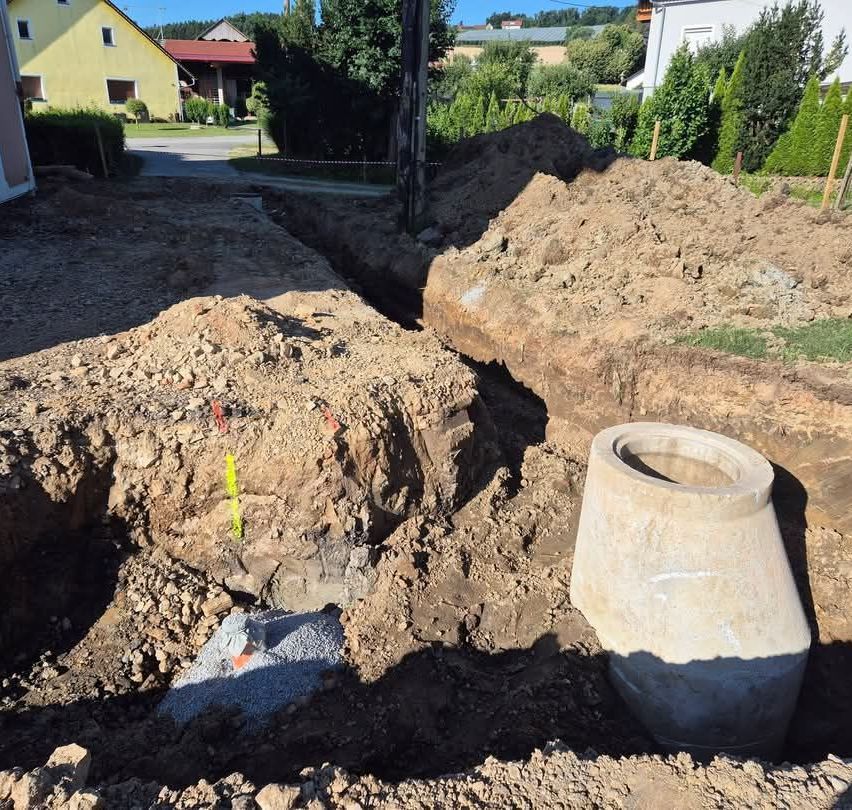 Eine Baustelle mit einem großen Betonrohr und Haufen von Erde und Kies. Ein gelbes Haus und Bäume sind im Hintergrund.