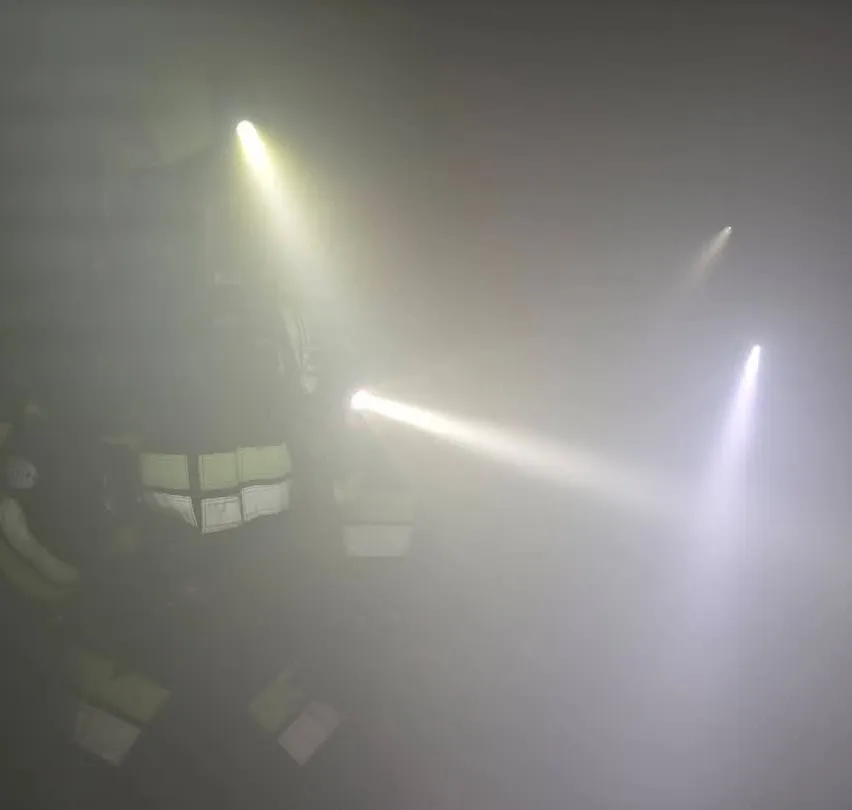 Ein Feuerwehrmann in voller Ausrüstung, beleuchtet von hellen Taschenlampen, steht in einer nebligen Umgebung.