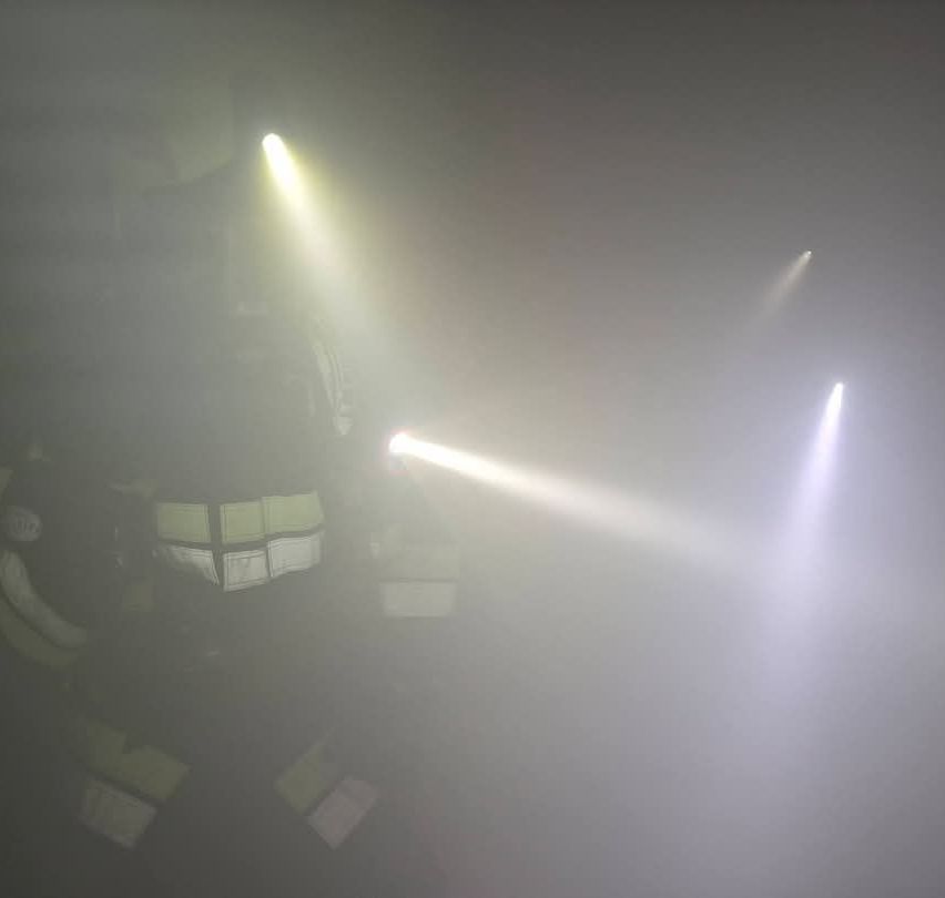 Ein Feuerwehrmann in voller Ausrüstung, beleuchtet von hellen Taschenlampen, steht in einer nebligen Umgebung.