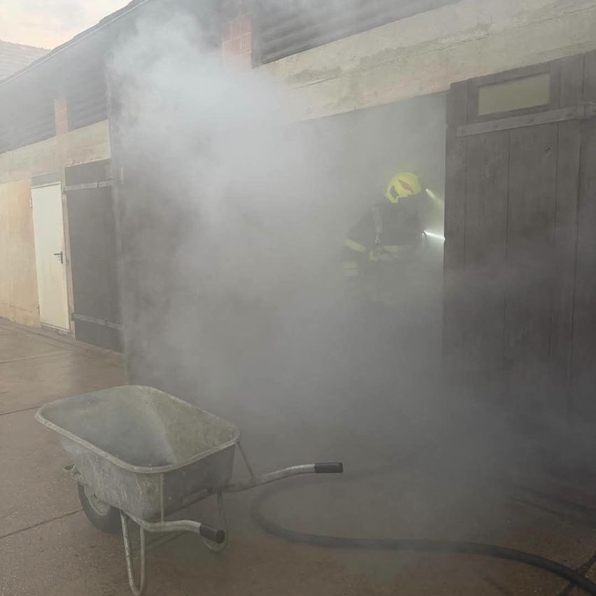 Ein Feuerwehrmann in voller Ausrüstung steht in einem rauchgefüllten Bereich, möglicherweise in einem Gebäude, mit einem Schubkarren und einem Schlauch in der Nähe.