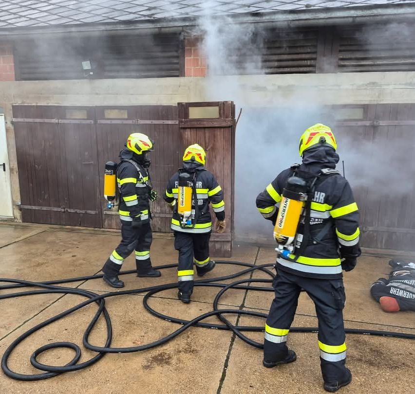 Drei Feuerwehrleute in voller Ausrüstung stehen vor einem Gebäude, aus dem Rauch aufsteigt. Sie halten Feuerlöscher und einer sprüht Wasser.