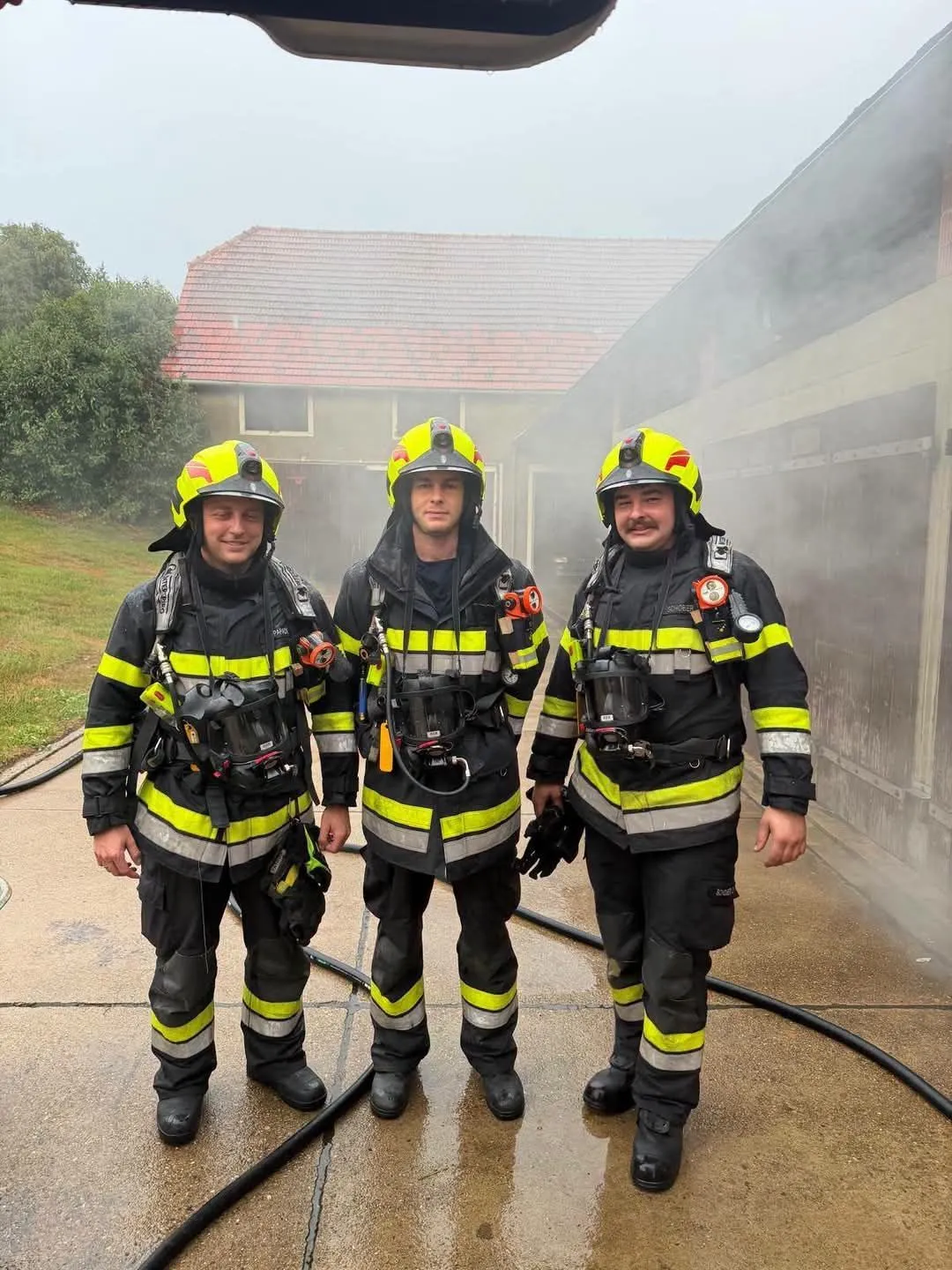 Drei Feuerwehrleute in voller Ausrüstung stehen vor einem Gebäude, aus dem Rauch aufsteigt. Das Gebäude hat ein rotes Dach und ist von Bäumen umgeben.