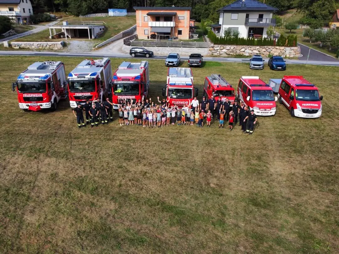 Eine Gruppe von Feuerwehrleuten und Kindern posiert für ein Foto mit mehreren Feuerwehrfahrzeugen im Hintergrund.