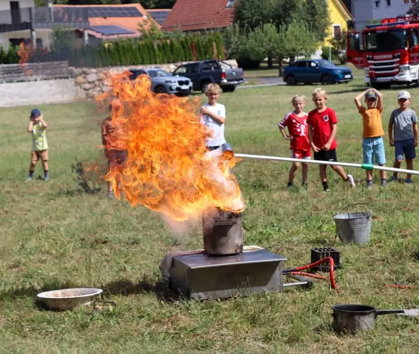 Kinder versammeln sich um einen Feuerkorb, aus dem Flammen aus einem Topf auf einem Metallständer aufsteigen, während andere aus der Ferne zusehen.