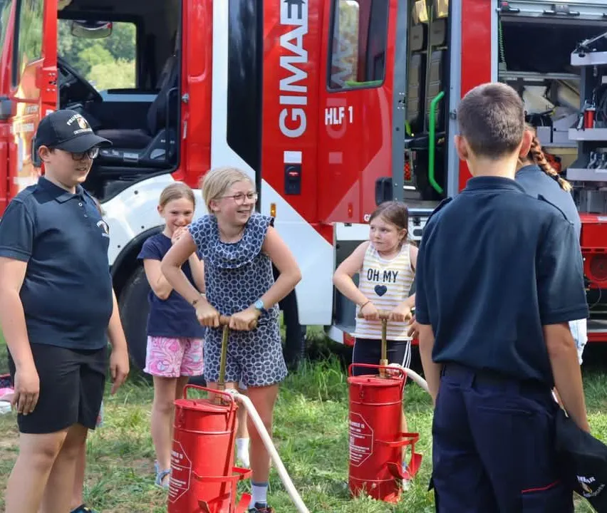 Eine Gruppe von Kindern und Erwachsenen steht um einen roten Feuerwehrwagen. Die Kinder halten rote Feuerlöscher.