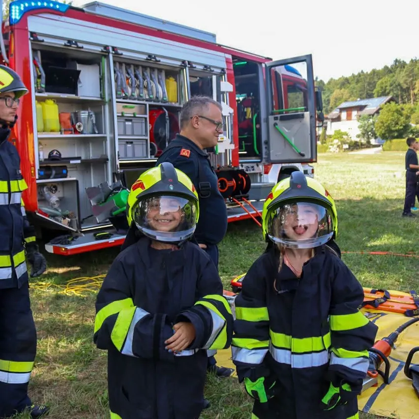 Zwei Kinder in Feuerwehruniformen stehen neben einem Feuerwehrauto mit offener Tür, während ein Mann in ähnlicher Uniform hinter ihnen steht.