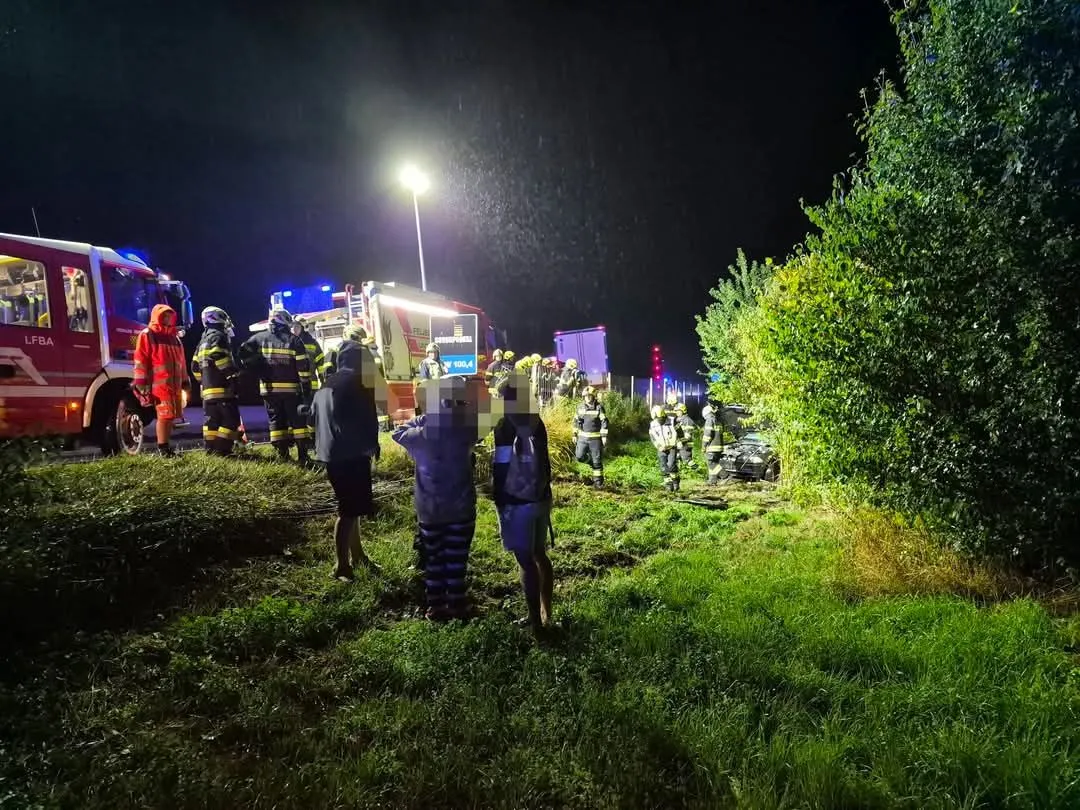 Notfallhelfer in Regenkleidung stehen neben einem demolierten Fahrzeug auf einem Rasen, mit einem Feuerwehrwagen und blinkenden Lichtern im Hintergrund.