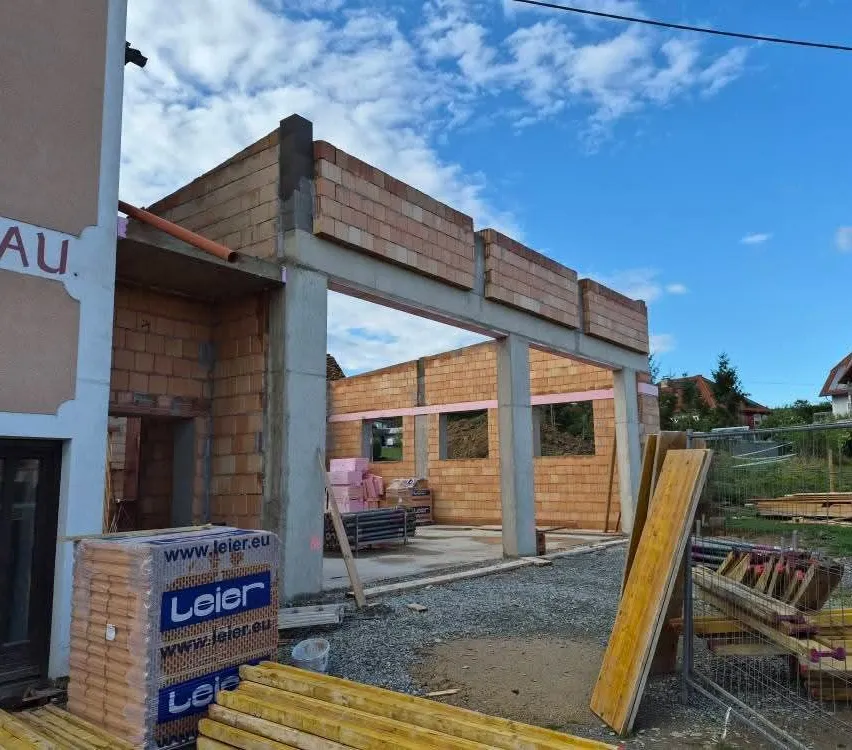 Eine Baustelle mit einem im Bau befindlichen Ziegelgebäude, einem klaren blauen Himmel im Hintergrund und verschiedenen Materialien und Werkzeugen, die herumliegen.