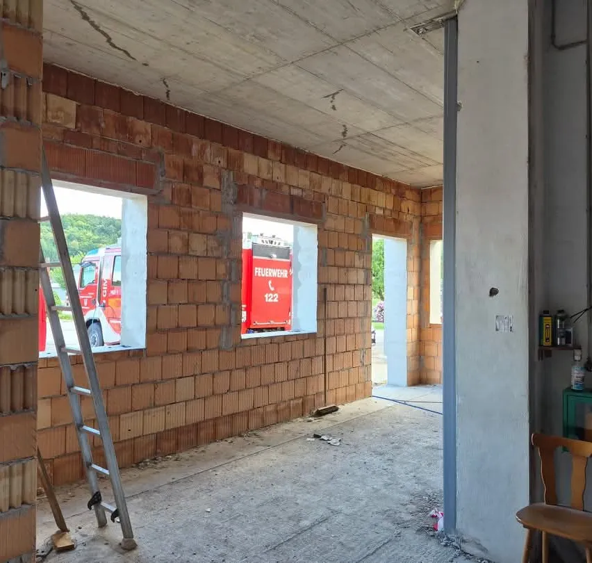 Ein Innenraum eines Gebäudes im Bau mit Ziegelwänden, einer Leiter und einem Feuerlöscher. Ein roter Feuerwehrwagen ist durch die Fenster draußen sichtbar.