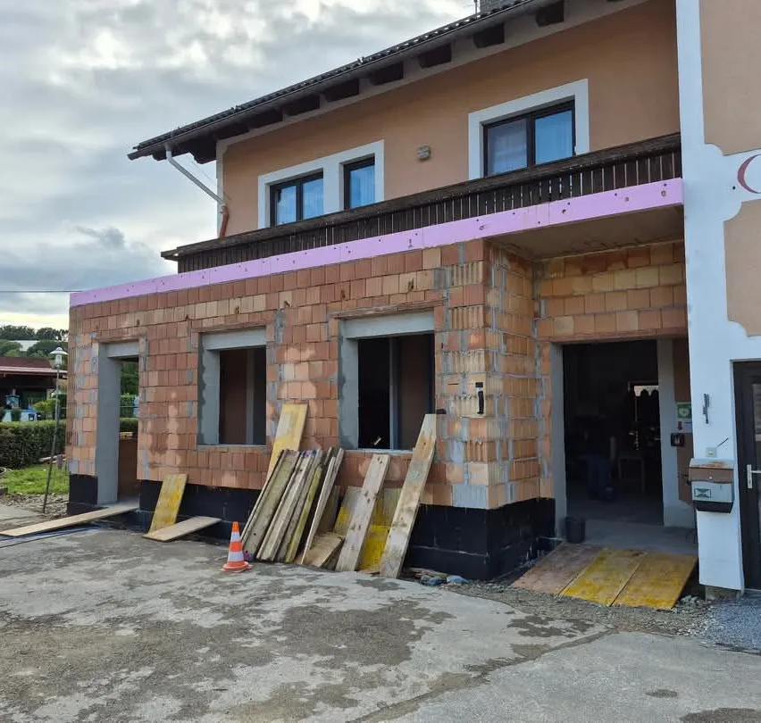 Ein Haus im Bau mit Ziegelwänden, einem Balkon und einer Garage. Holzbretter und ein orangefarbener Verkehrskegel sind in der Nähe.