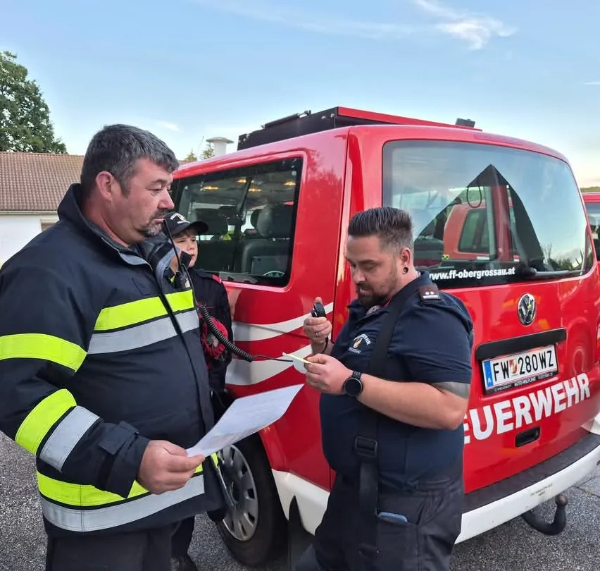 Zwei Feuerwehrleute stehen vor einem roten Fahrzeug, einer hält ein Papier und der andere ein Funkgerät. Ein Kind ist im Fahrzeug.