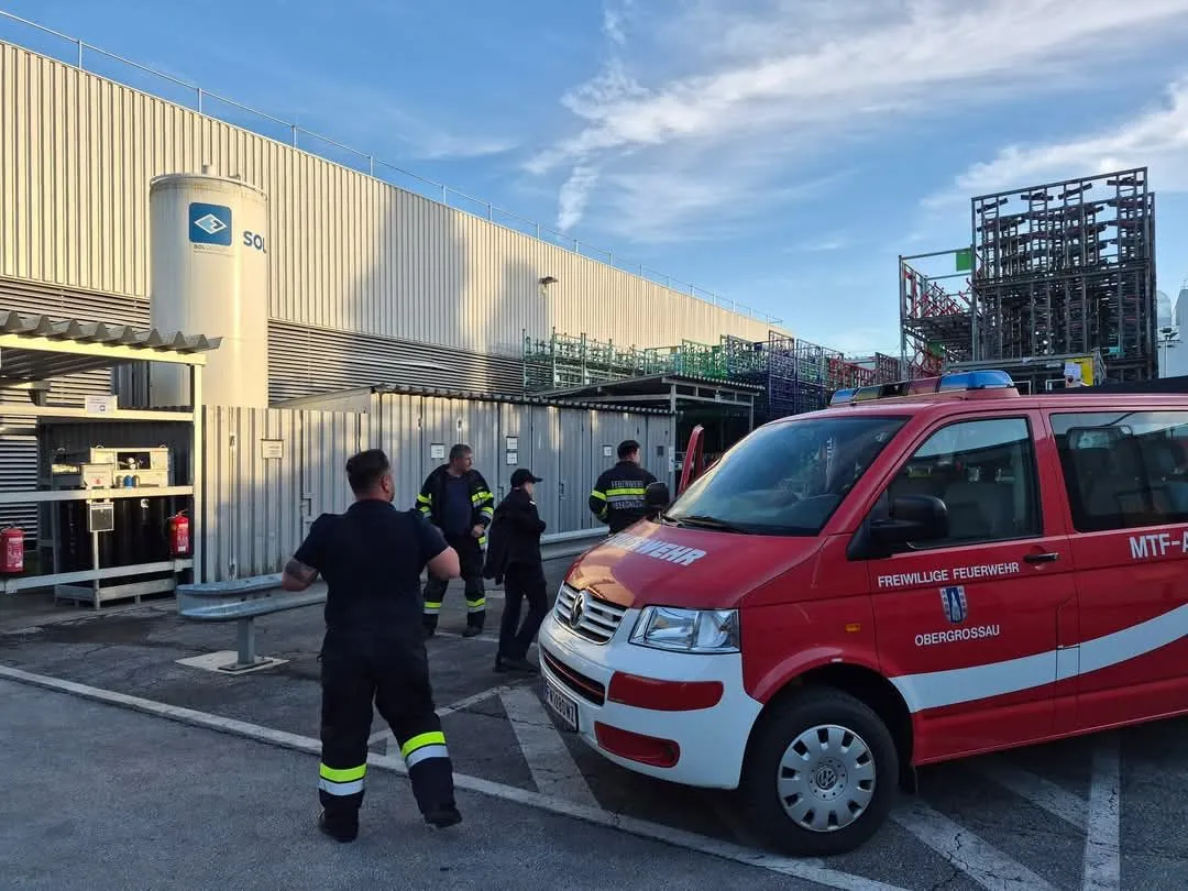 Ein roter Feuerwehrwagen ist vor einem Gebäude mit einem großen weißen Tank geparkt. Mehrere Feuerwehrleute stehen um den Wagen herum.