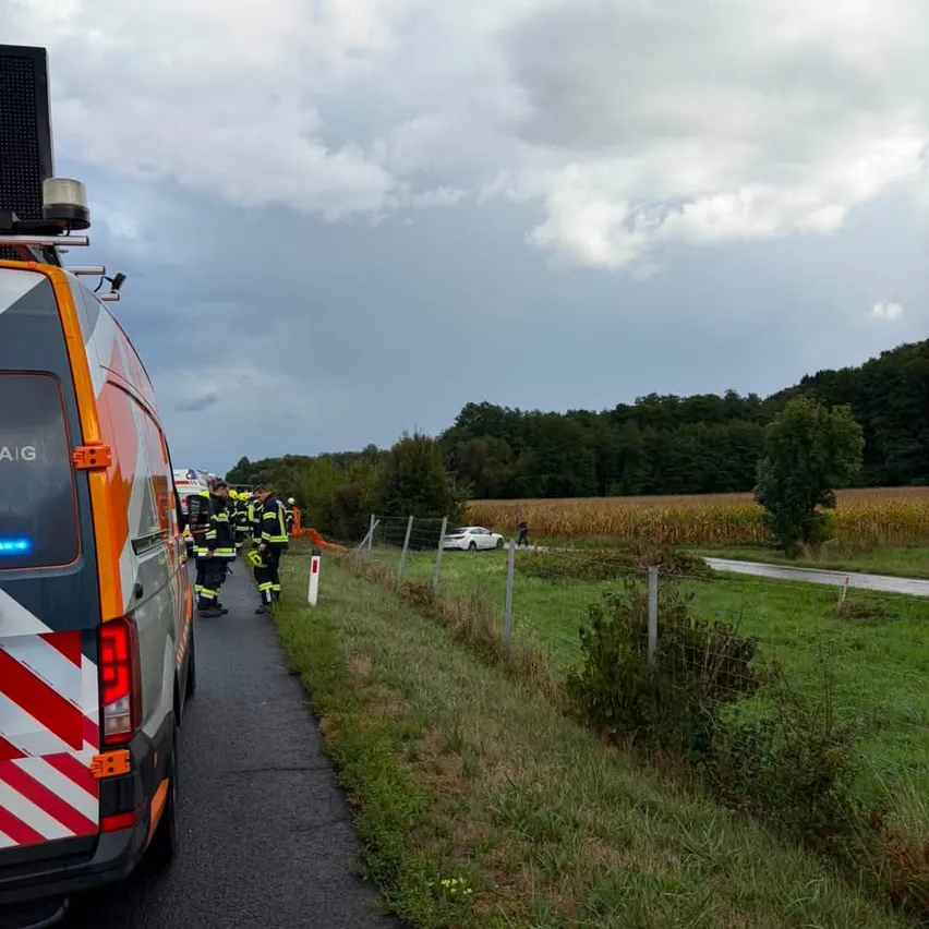 Ein Rettungsteam kümmert sich um einen Verkehrsunfall auf einer ländlichen Straße, mit einem weißen Auto am Straßenrand und einem bewölkten Himmel.