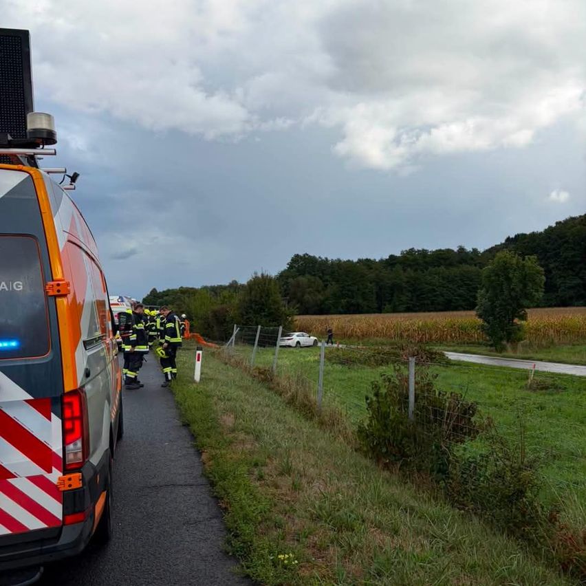 Ein Rettungsteam kümmert sich um einen Verkehrsunfall auf einer ländlichen Straße, mit einem weißen Auto am Straßenrand und einem bewölkten Himmel.