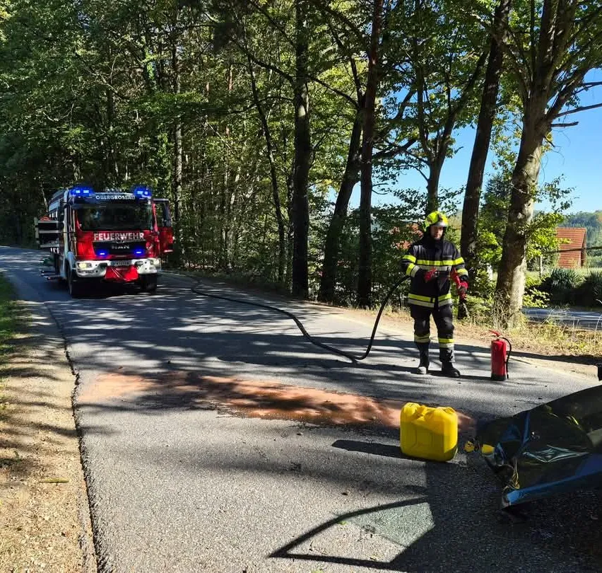 Ein Feuerwehrmann steht am Straßenrand, hält einen Schlauch und sprüht Wasser auf einen Feuerlöscher.
