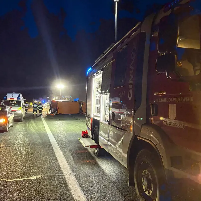 Ein Feuerwehrauto ist nachts am Straßenrand geparkt. Es gibt mehrere Rettungsfahrzeuge und Menschen in der Nähe, die möglicherweise einen Unfall bearbeiten.