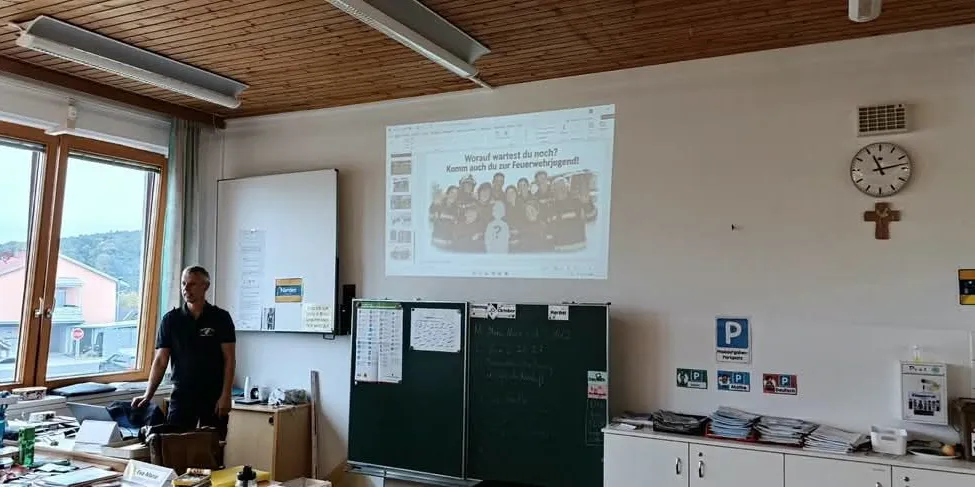 Ein Mann steht in einem Klassenzimmer mit einer Projektionsleinwand, die ein Gruppenfoto zeigt.