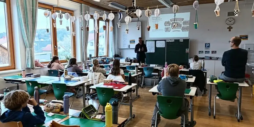 Ein Klassenzimmer mit Schülern an Schreibtischen, einem Lehrer, der vor einer Projektionsleinwand steht, und einer Uhr an der Wand.
