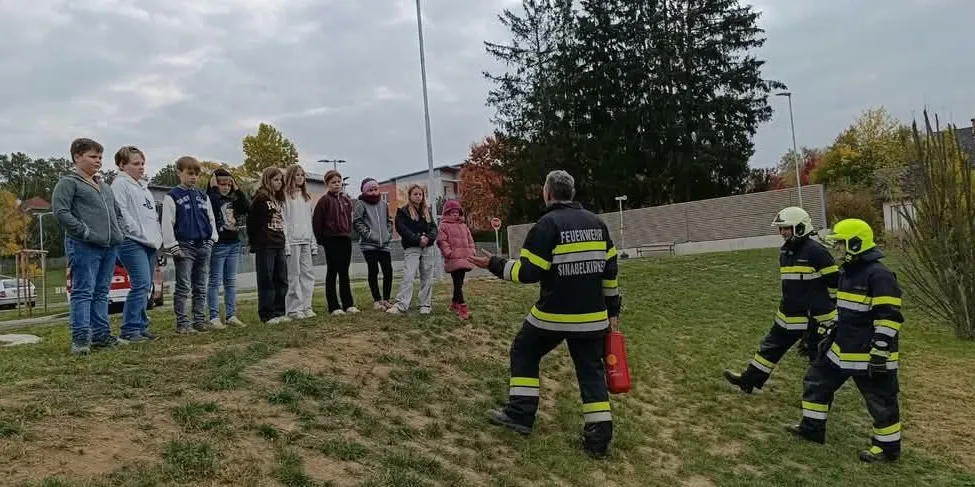 Eine Gruppe von Menschen, einschließlich Kindern, steht in einer Reihe auf einem Grasfeld. Zwei Feuerwehrleute demonstrieren ihnen etwas.