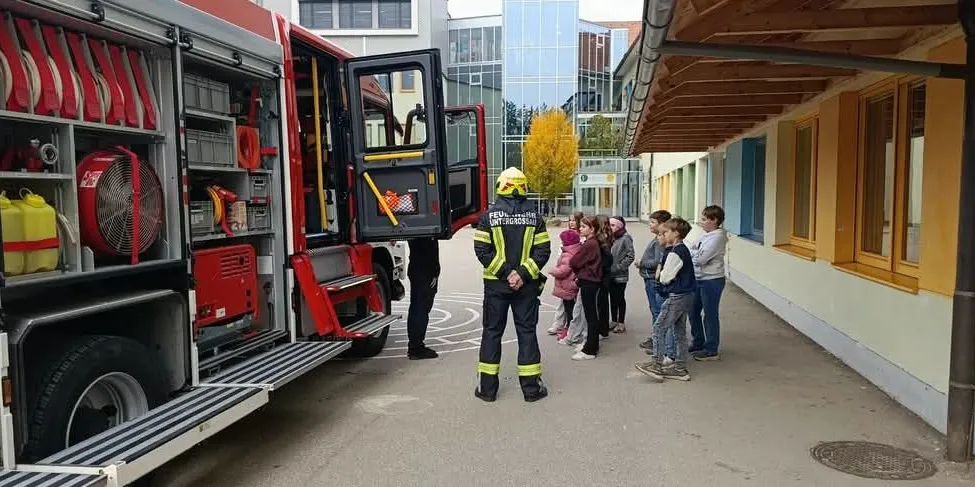 Ein Feuerwehrmann steht neben einem Feuerwehrauto mit offener Tür. Eine Gruppe von Kindern steht vor ihm und schaut zu.