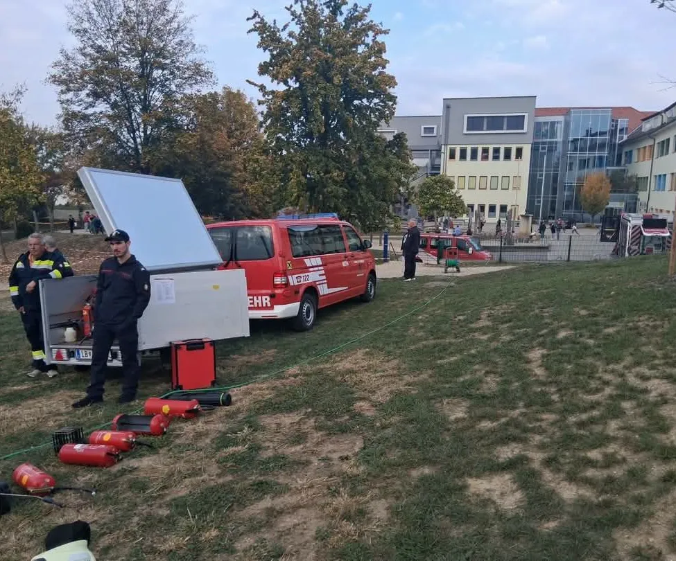 Ein rotes Rettungsfahrzeug mit offener Hecktür steht auf einem Rasenfeld. Zwei Personen stehen neben dem Fahrzeug, und mehrere Feuerlöscher liegen auf dem Boden.