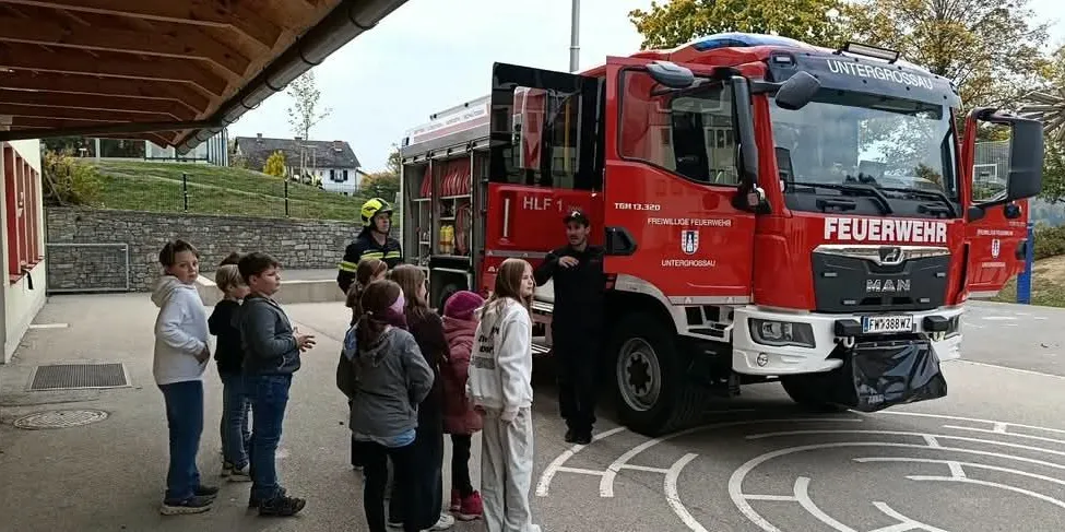 Eine Gruppe von Kindern steht um einen roten Feuerwehrwagen herum, vor dem zwei Feuerwehrleute stehen.
