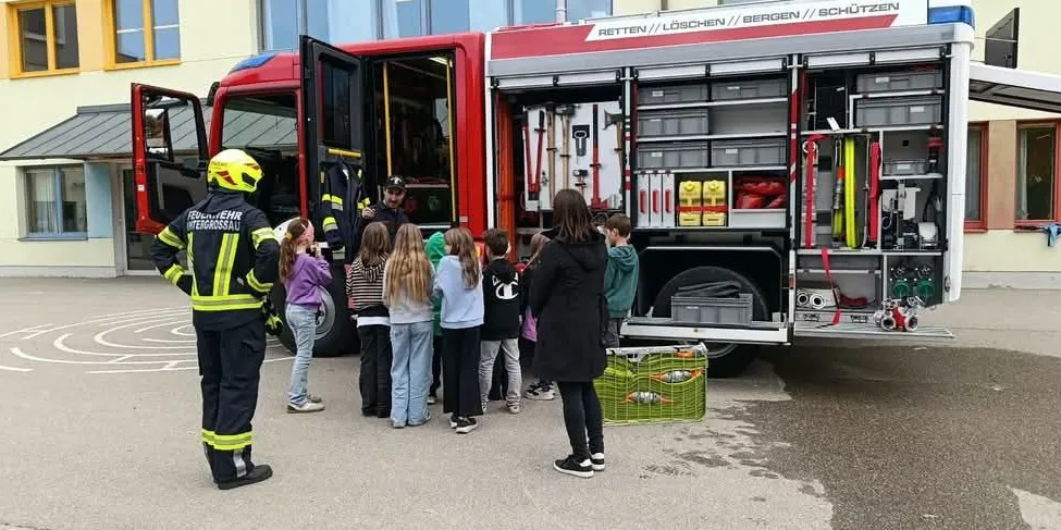 Eine Gruppe von Kindern steht um einen Feuerwehrwagen herum, neben ihnen steht ein Feuerwehrmann.