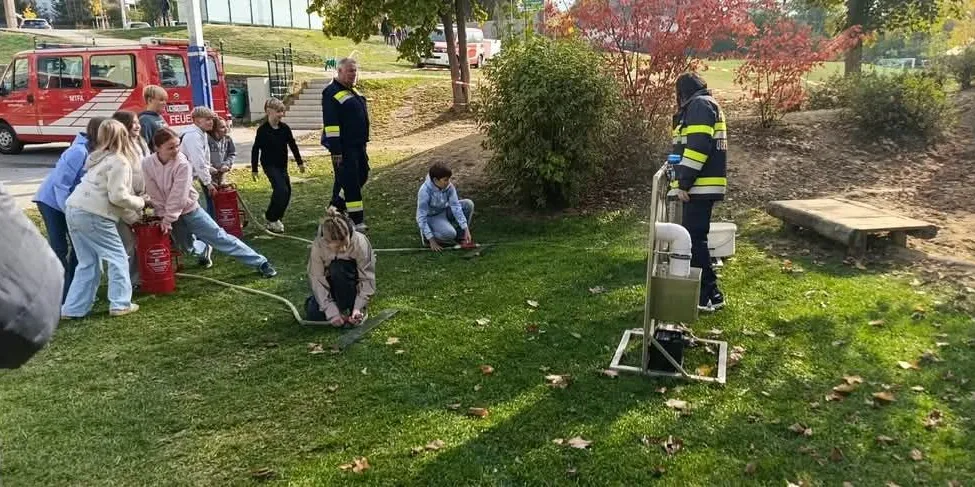 Eine Gruppe von Menschen, einschließlich Kindern, versammelt sich auf einem Rasenfeld. Sie halten rote Feuerlöscher und scheinen Brandschutzübungen zu machen. Ein Feuerwehrmann steht in der Nähe und hält einen Schlauch.