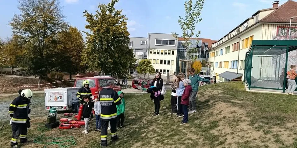 Eine Gruppe von Kindern und Erwachsenen steht auf einem Rasenfeld, wahrscheinlich in einem Park. Sie tragen Schutzausrüstung und nehmen möglicherweise an einer Brandschutzübung teil. Ein Feuerwehrauto und ein Feuerlöscher liegen auf dem Boden.