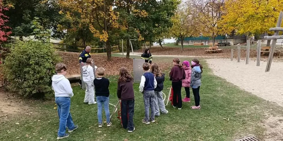 Eine Gruppe von Kindern steht im Kreis in einem Park mit Bäumen und einem Spielplatz im Hintergrund.