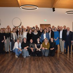 Eine Gruppe von Menschen versammelt sich für ein Foto in einem Raum mit Holzboden. Einige stehen, andere sitzen. Ein Schild mit dem Text 'STEHT ALLEN' ist an der Wand angebracht.