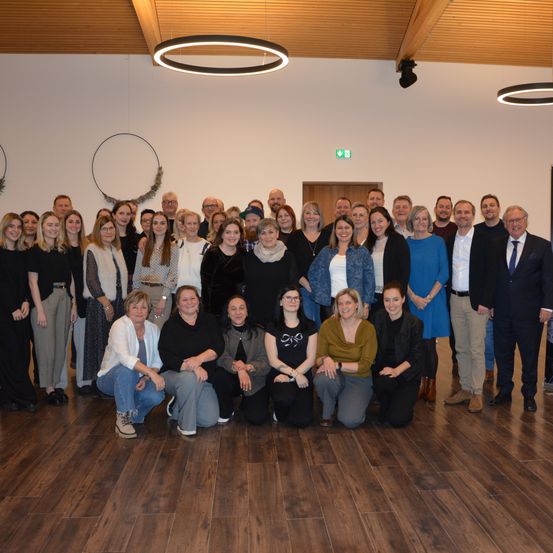 Eine Gruppe von Menschen versammelt sich für ein Foto in einem Raum mit Holzboden. Einige stehen, andere sitzen. Ein Schild mit dem Text 'STEHT ALLEN' ist an der Wand angebracht.