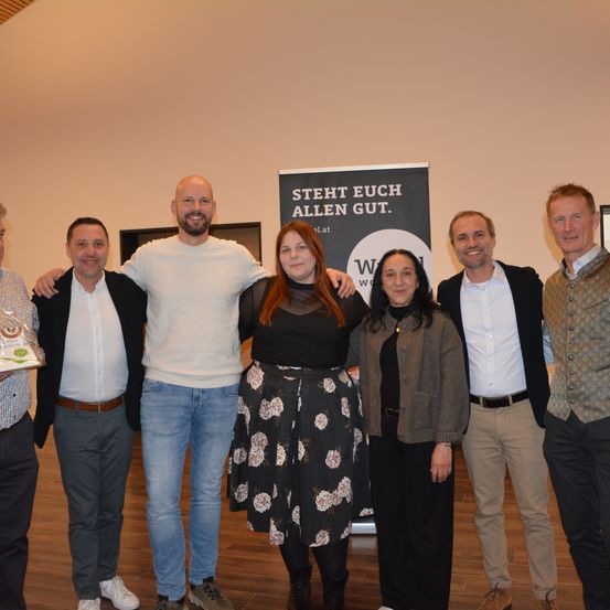 Eine Gruppe von Menschen posiert für ein Foto mit einem Banner, das 'Bleibt allen gut' sagt. Dahinter ist eine weiße Wand.