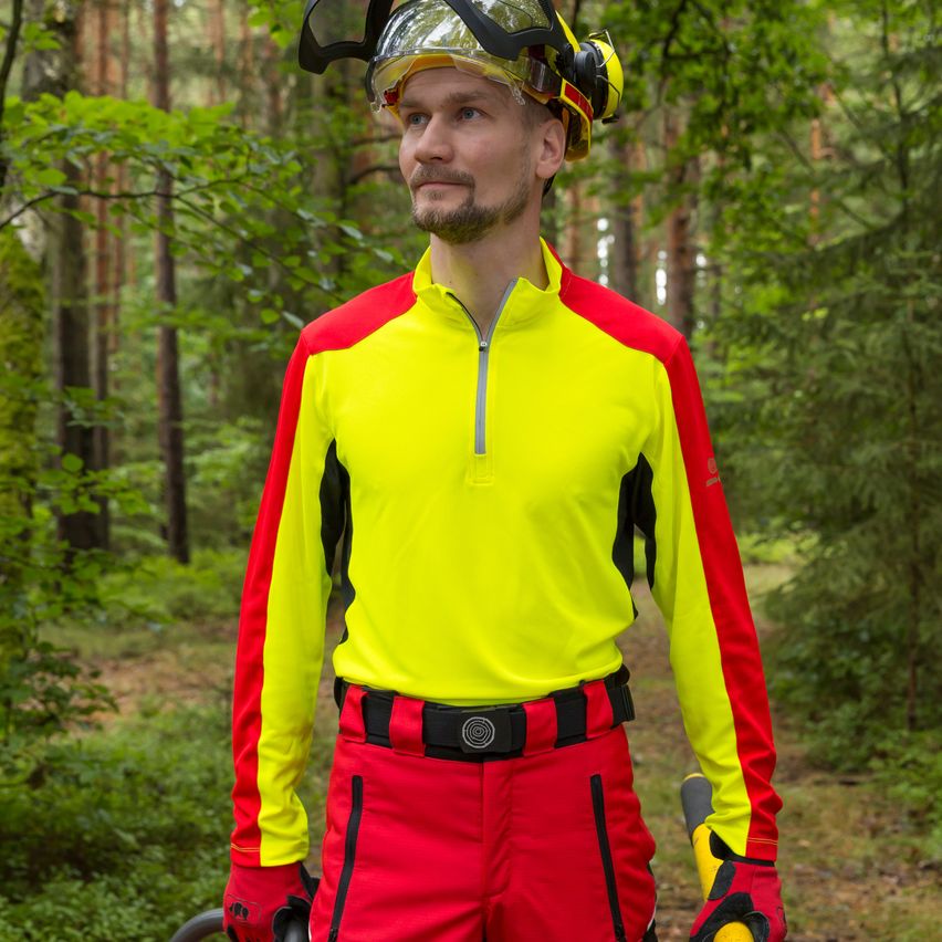 A man in bright yellow and red high-visibility clothing stands in a forest, wearing a helmet and gloves. He is looking to the side, possibly posing for a photo.