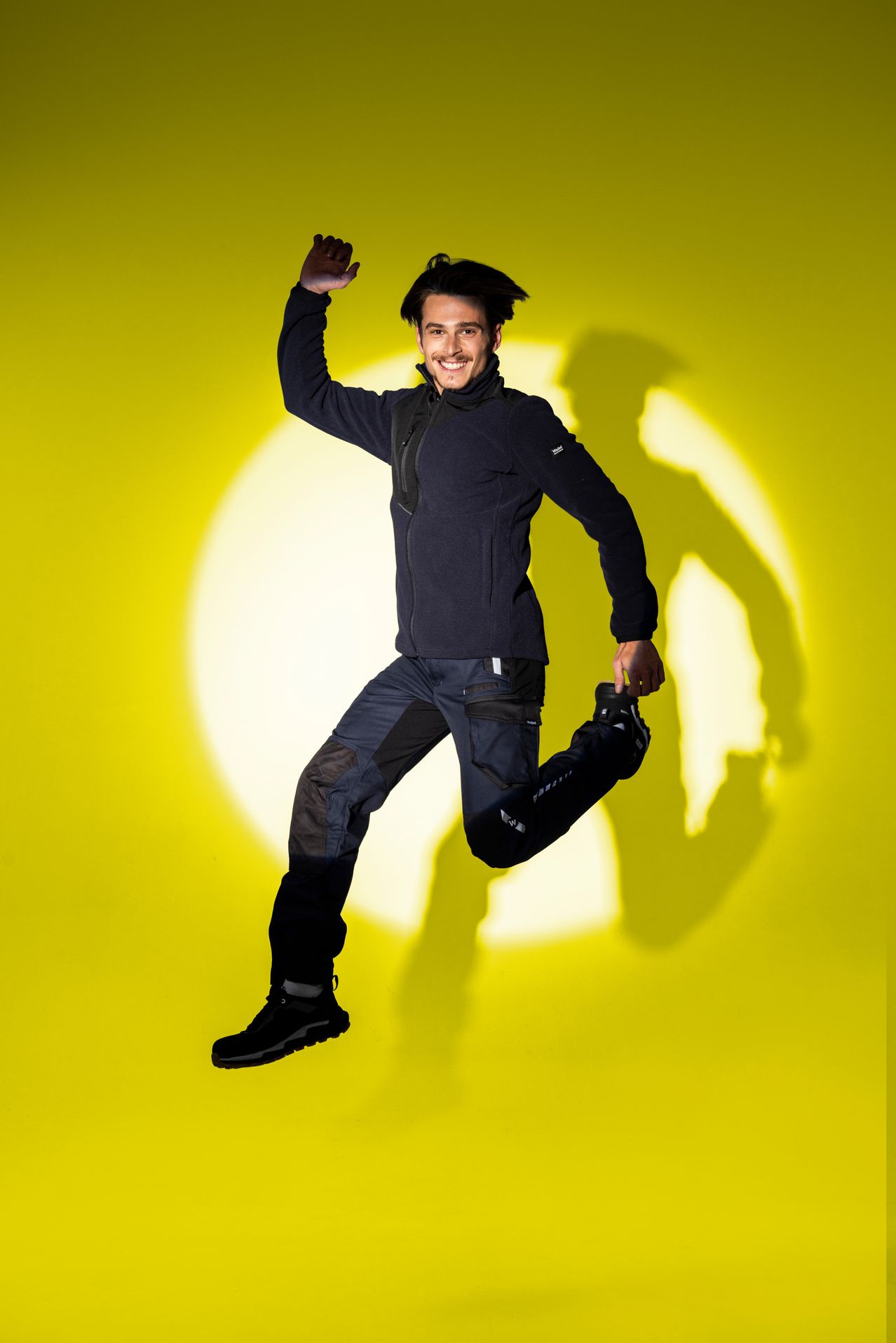 Ein Mann springt mit einem erhobenen Bein, er trägt eine schwarze Jacke und Hose vor einem leuchtend gelben Hintergrund.