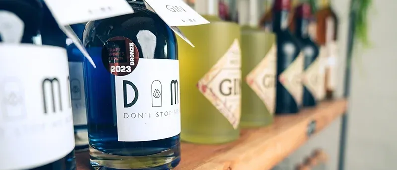 Ein Holzregal beherbergt eine Reihe von Flaschen, darunter eine blaue mit der Aufschrift '2023' und 'Don't Stop Me', zusammen mit anderen, die Etiketten wie 'GIN' und 'G' tragen.