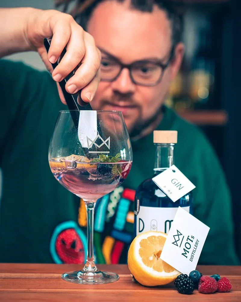 Ein Barkeeper mit Brille mischt ein Getränk in einem hohen Glas. Das Getränk ist mit Zitrone und Minze garniert. Neben dem Glas steht eine Flasche mit der Aufschrift 'Gin' und eine weitere Flasche mit der Aufschrift 'Moots Distillery'.