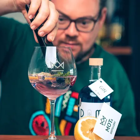 Ein Barkeeper mit Brille mischt ein Getränk in einem hohen Glas. Das Getränk ist mit Zitrone und Minze garniert. Neben dem Glas steht eine Flasche mit der Aufschrift 'Gin' und eine weitere Flasche mit der Aufschrift 'Moots Distillery'.
