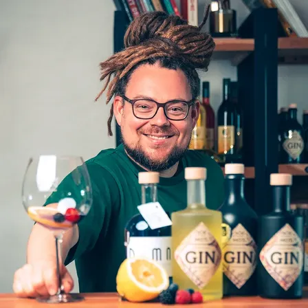 Ein lächelnder Mann mit Dreadlocks, Brille und grünem Shirt sitzt hinter einer Bar mit verschiedenen Flaschen und einem Weinglas. Er hält ein Weinglas mit Früchten und ist von Ginflaschen umgeben.