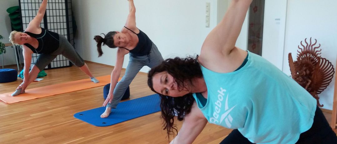 Drei Frauen in einem Yogastudio führen Yoga-Posen auf orangefarbenen Matten durch. Eine Frau in einem türkisen Shirt lächelt in die Kamera.