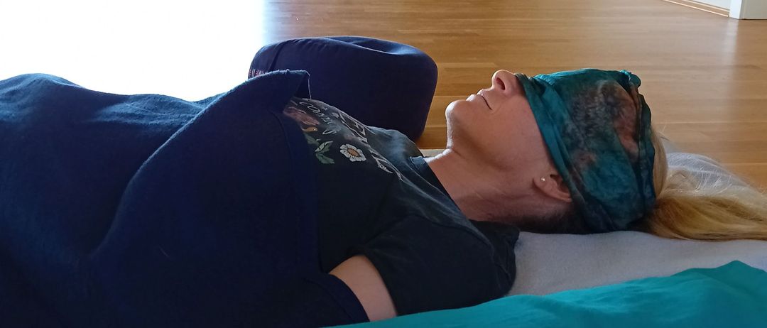 Eine Frau liegt mit geschlossenen Augen auf einem Bett. Ein türkiser Polster ist neben ihr, und eine Pflanze ist im Hintergrund.