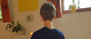 Eine Person in einem blauen Umhang sitzt in einem Raum und schaut vom Kamera weg. Hinter ihr steht ein Tisch mit einer Kerze und einer Pflanze. Ein Bilderrahmen und ein gelbes Gemälde sind an der Wand. Ein Topf mit einer Pflanze steht auf der Fensterbank.