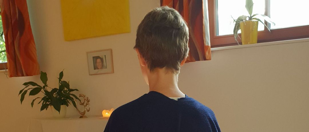 Eine Person in einem blauen Umhang sitzt in einem Raum und schaut vom Kamera weg. Hinter ihr steht ein Tisch mit einer Kerze und einer Pflanze. Ein Bilderrahmen und ein gelbes Gemälde sind an der Wand. Ein Topf mit einer Pflanze steht auf der Fensterbank.