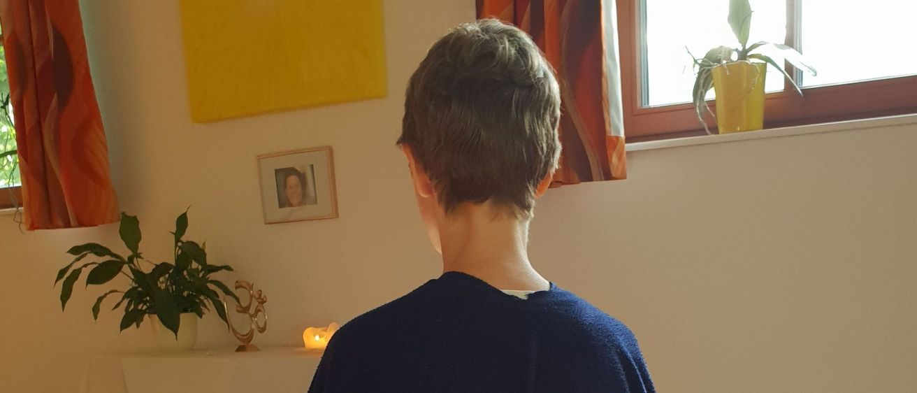 Eine Person in einem blauen Umhang sitzt in einem Raum und schaut vom Kamera weg. Hinter ihr steht ein Tisch mit einer Kerze und einer Pflanze. Ein Bilderrahmen und ein gelbes Gemälde sind an der Wand. Ein Topf mit einer Pflanze steht auf der Fensterbank.