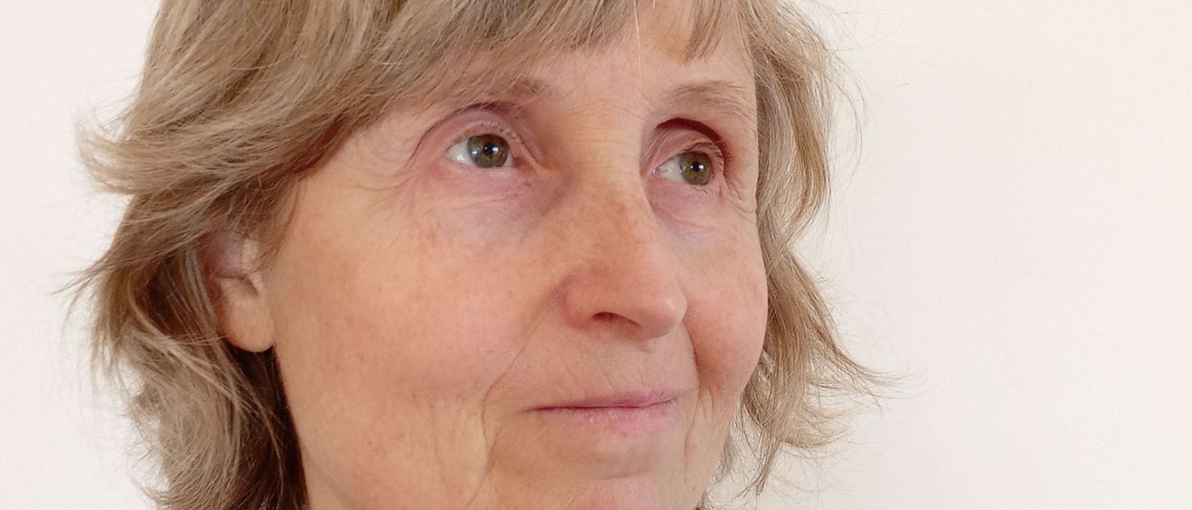 Eine Frau mit kurzen, welligen blonden Haaren schaut direkt in die Kamera. Sie trägt eine hellblaue Jacke über einem weißen Hemd und hat Falten um die Augen und den Mund.