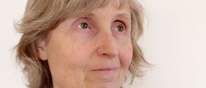 Eine Frau mit kurzen, welligen blonden Haaren schaut direkt in die Kamera. Sie trägt eine hellblaue Jacke über einem weißen Hemd und hat Falten um die Augen und den Mund.